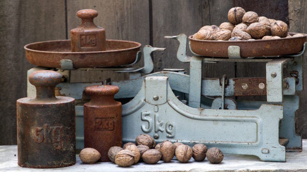 Eine alte Balkenwaage. Auf der einen Seite befindet sich 1 Kilogramm, auf der anderen Walnüsse. Weitere Gewichte und Walnüsse sind im Vordergrund zu sehen.