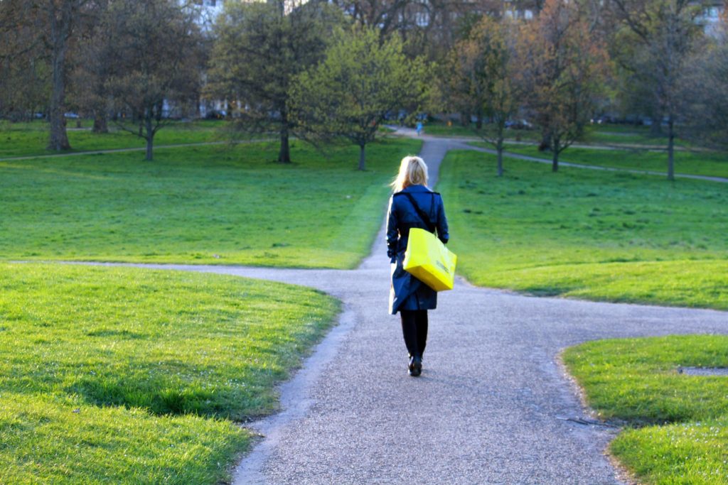 Eine blonde Frau, die durch einen Park läuft. Sie trägt eine gelbe Tasche und nähert sich einer Weggabelung.