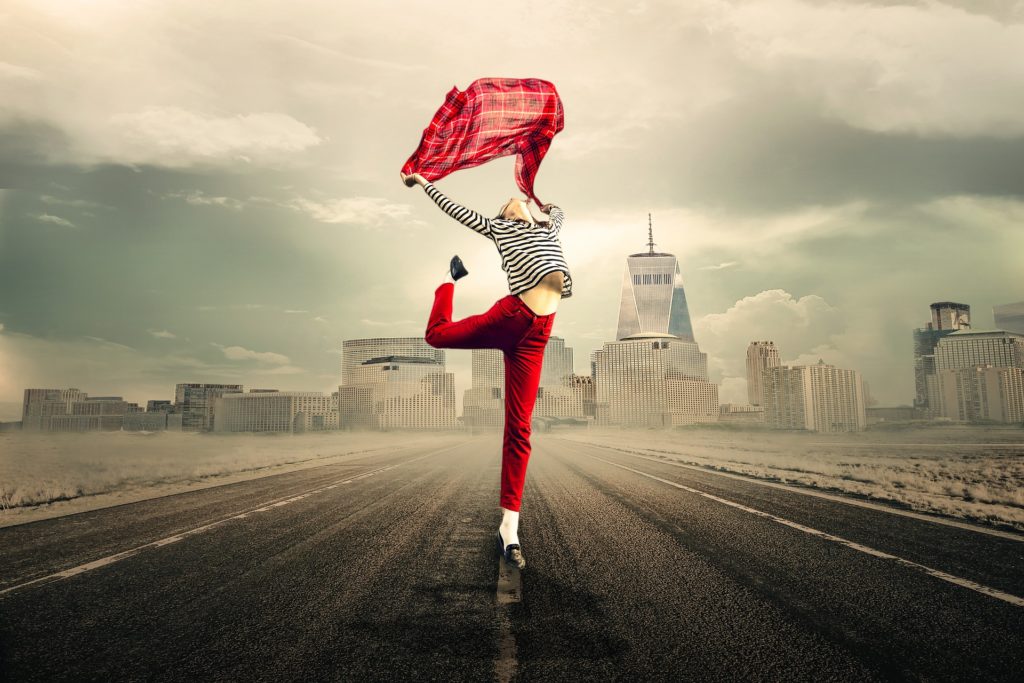 Eine Frau mit roter Hose, gestreiften Shirt und einem rot-karierten Tuch tanzt auf einer leeren Straße.