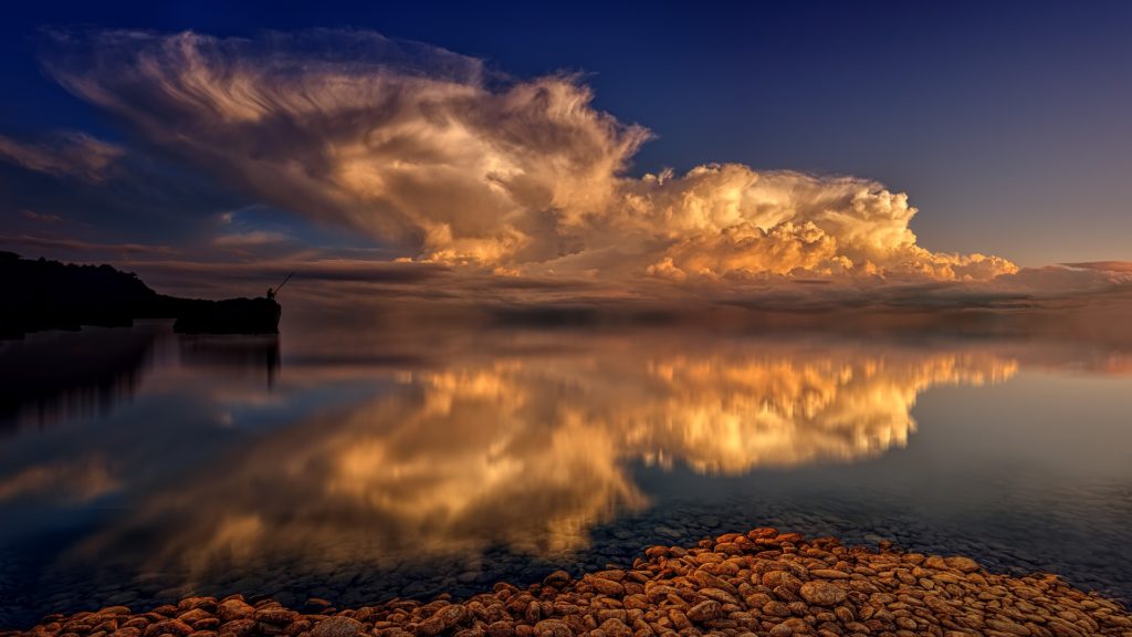Ein spiegelglatter See, an dessen Ufer ein Angler sitzt. Die aufgehende Sonne beleuchtet Wolken, die sich im Wasser spiegeln